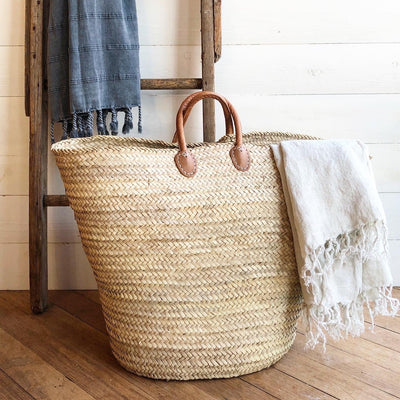 French laundry market tote displayed with vintage wooden latter and white linens. Tote is light tan with cognac brown leather handles. 