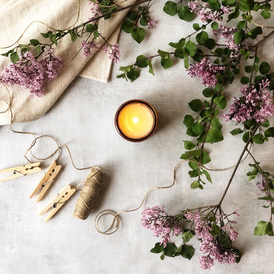 Flat lay for candle in the scent Sunday. Features burning candle in amber glass jar, lilac branches, spool of twine, and wooden clothespins. 