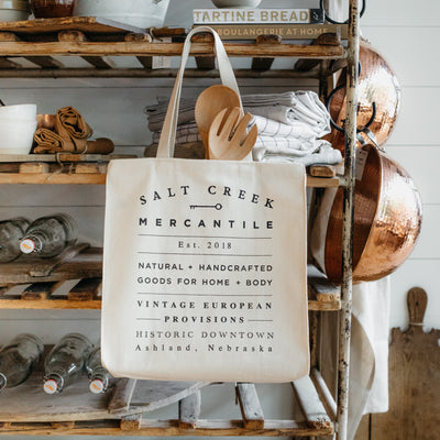 Natural canvas tote with Salt Creek Mercantile logo. Displayed on a vintage wooden rack with linens, copper bowls, wooden boards, serveware, and glass bottles.  