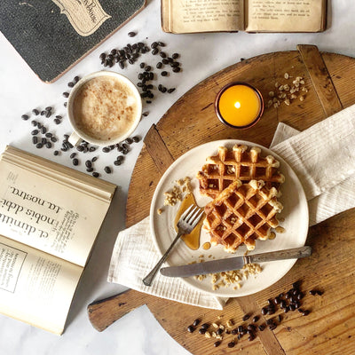 Lit coffee shop candle in amber glass jar displayed on vintage wooden board with waffles and syrup, open books, a foamy latte and coffee beans. 