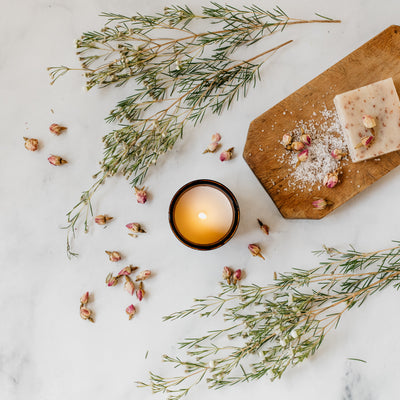 Flat lay of Caroline candle featuring burning candle in amber glass jar, handmade floral soap, green herb sprigs, and dried florals. 