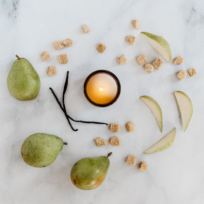 Flat lay photo of candle in the scent Vintage Pear. Features green pears, raw sugar cubes, vanilla bean, and burning candle in amber glass jar. 