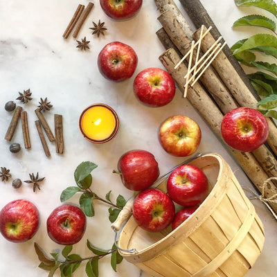 Lit orchard candle displayed with red apples toppling out of a wooden basket, cinnamon sticks, whole cloves, wooden branches, and green leaves. 